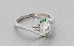 Contemporary Bypass Design Engagement Ring Set With A Round Brilliant Diamond And Framed With Forest Green Emeralds.