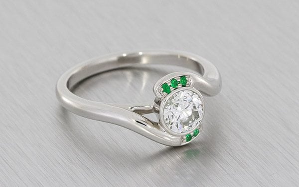 Contemporary Bypass Design Engagement Ring Set With A Round Brilliant Diamond And Framed With Forest Green Emeralds.